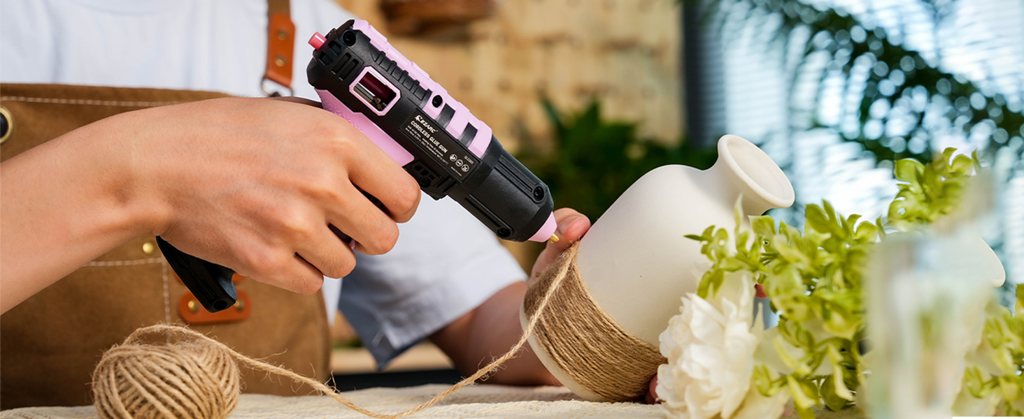 Cordless glue gun being used on crafting materials, with twine and decorative plants visible in the background.