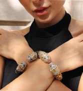 Close-up of hands wearing two ornate gold bracelets with intricate designs and gemstone accents against a black top.