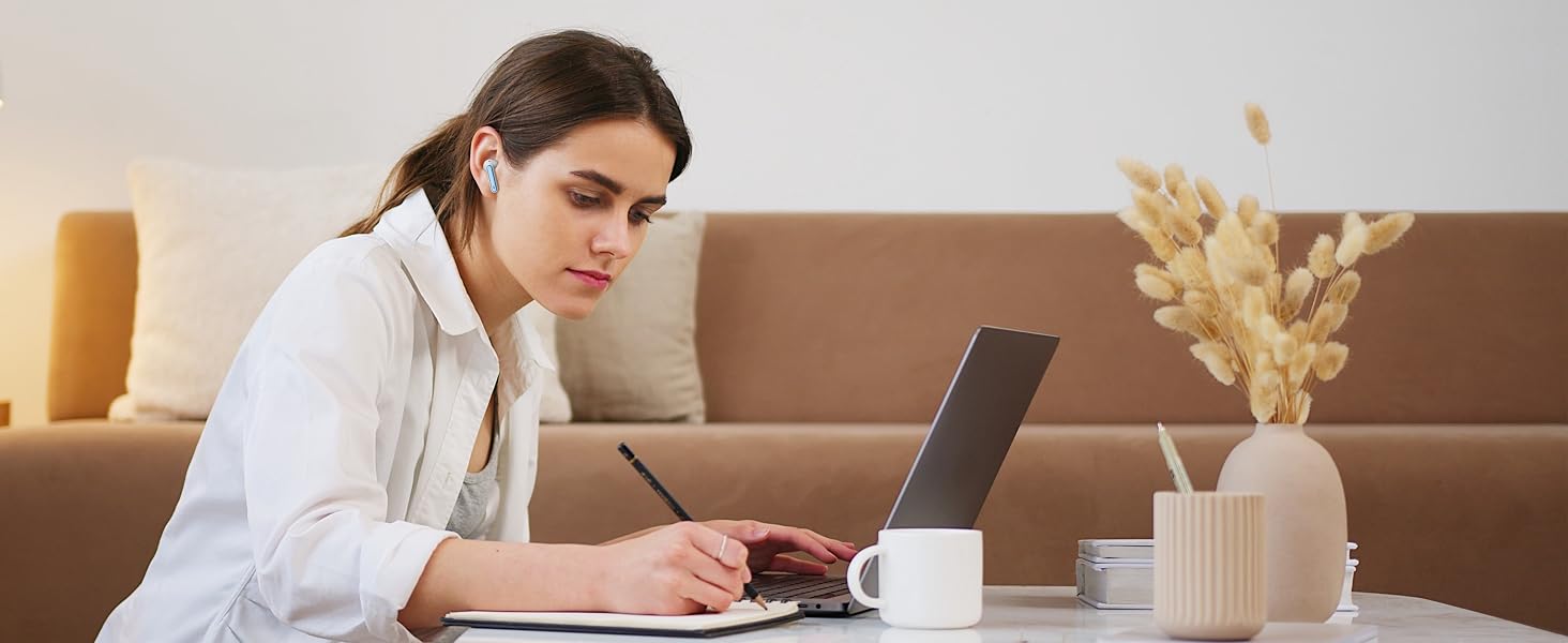 Woman using laptop on coffee table in living room. White mug, decorative vase with dried flowers, and textured candle holder visible on table.