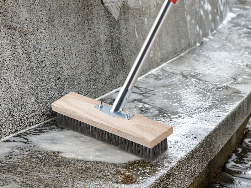 Push broom with wooden block head and stiff bristles cleaning wet concrete surface. Long metal handle attached to broom head. Water visible on concrete.