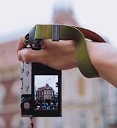 Camera with olive green wrist strap being held while capturing architectural photo, showing camera screen with building in frame.