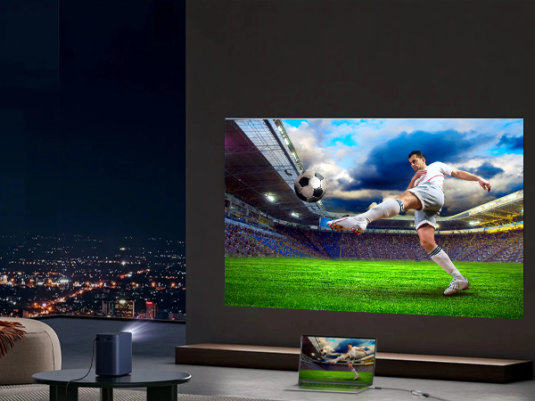 Large screen displaying soccer player kicking ball in stadium. Shown in dimly lit room with city view through window.