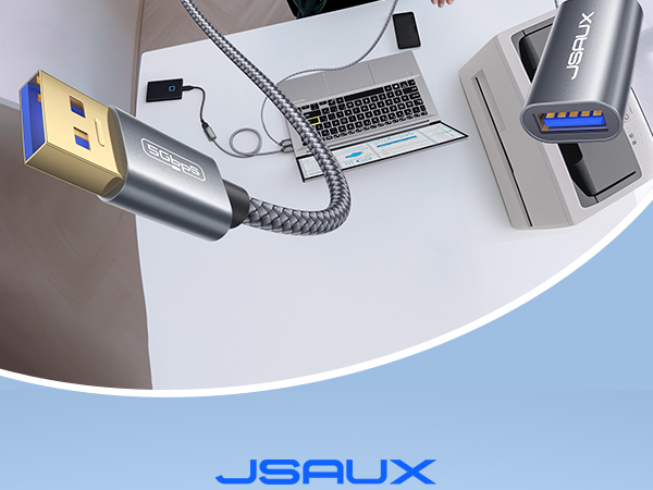 USB cable with gold-plated connector and braided cord, connected to a laptop. Portable battery pack with LED indicators visible nearby. JSAUX brand logo at bottom.