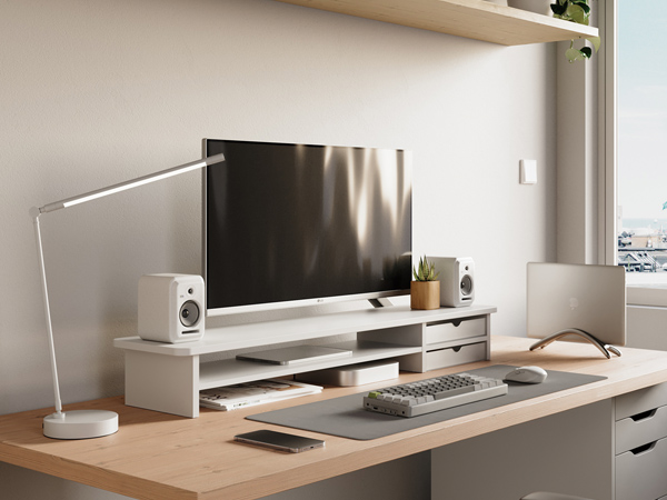 Modern home office setup with white desk, computer monitor, keyboard, mouse, desk lamp, and wall-mounted shelving unit against a white wall.