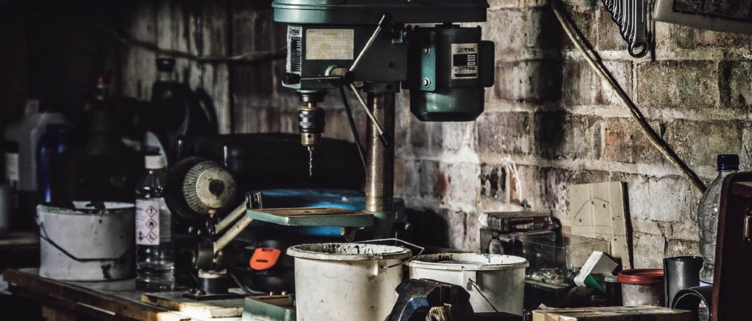 Industrial workshop setting with various tools and equipment on a metal workbench, including drill press and assorted containers against a brick wall background.