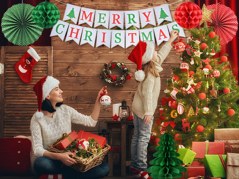 Christmas scene with decorated tree, 'Merry Christmas' banner, and festive decor. Two people arranging ornaments and decorations in a cozy room.