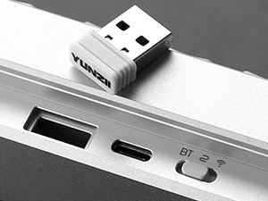 Small white USB adapter plugged into a laptop port. The laptop's side shows multiple ports including USB-C and a switch labeled 'BT'.