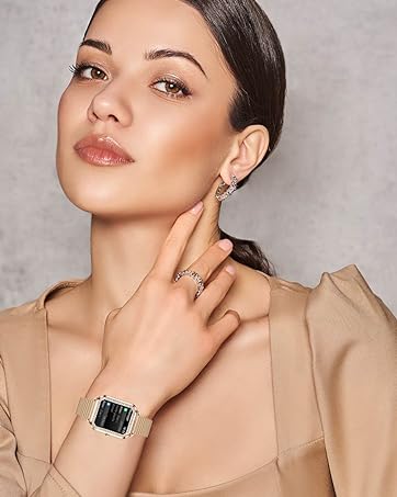 Woman wearing beige top, touching her face. She has dark hair and is wearing makeup. A rectangular watch is visible on her wrist.