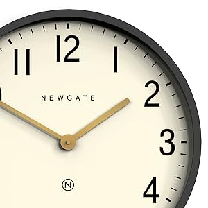 Newgate wall clock with black frame, cream-colored dial, and bold black numerals. Features brass-colored hands and modern minimalist design.