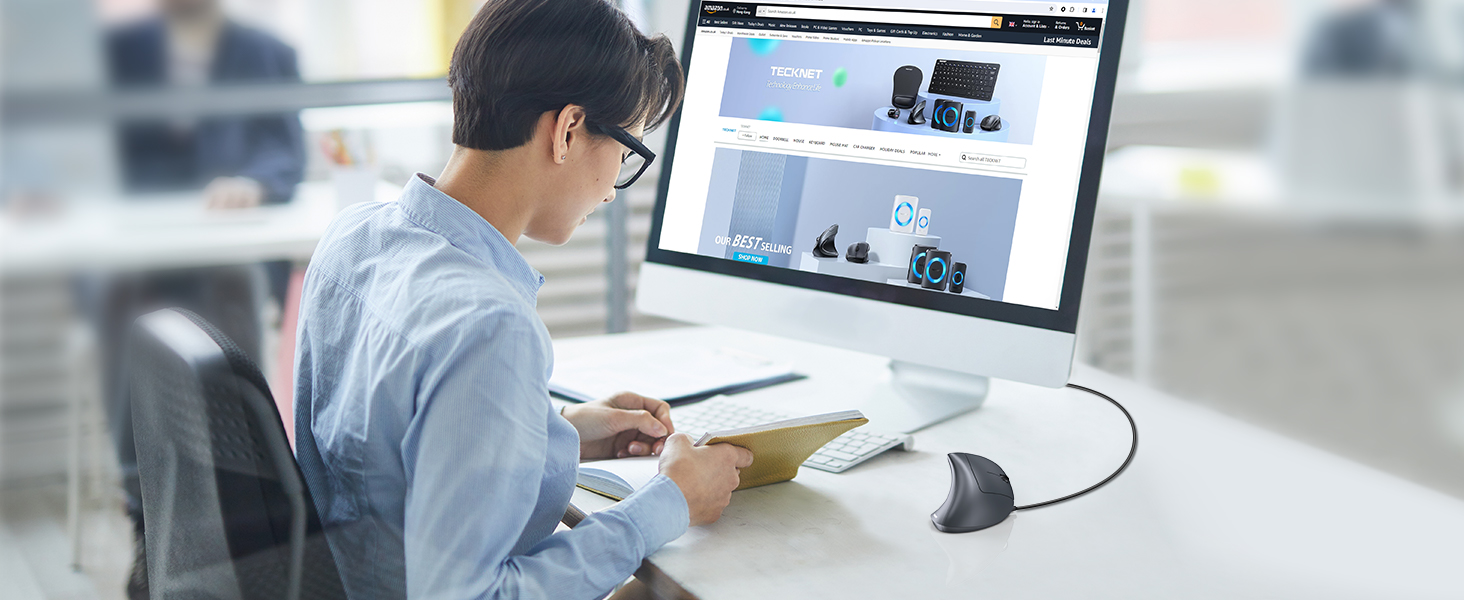 Person working at desk with computer monitor displaying a website, using a mouse and reviewing documents in an office setting.