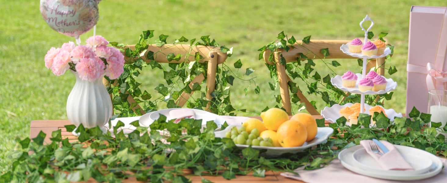 Outdoor picnic setup on grass featuring pink floral arrangement, decorative items, and food display on checkered cloth with residential building background.