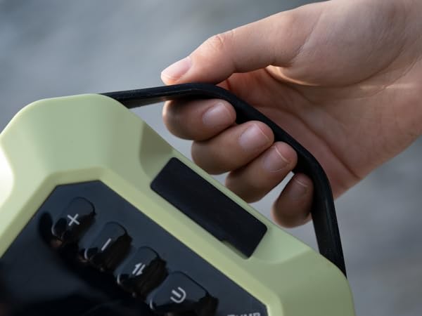 Hand holding a pale green electronic device with black buttons and a carrying handle. The device appears to be a portable control unit or measurement tool.