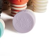 Text reads 'Colo Clean'. Close-up product photo showing light purple circular soap or cleaning product with embossed text on its surface, surrounded by stacked colorful soaps.