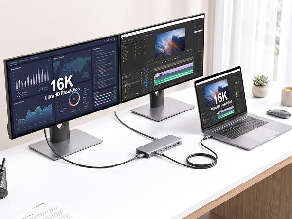 Text reads '16K'. Dual monitor desktop setup with two displays showing data visualizations, silver monitor stand, laptop, white accessories, desk lamp, and cables on wooden desk.