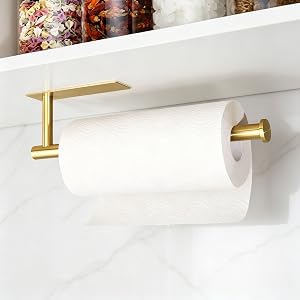Gold-finish paper towel holder mounted on white wall. Horizontal bar design with decorative floral arrangement visible in background. Modern minimalist style fixture.