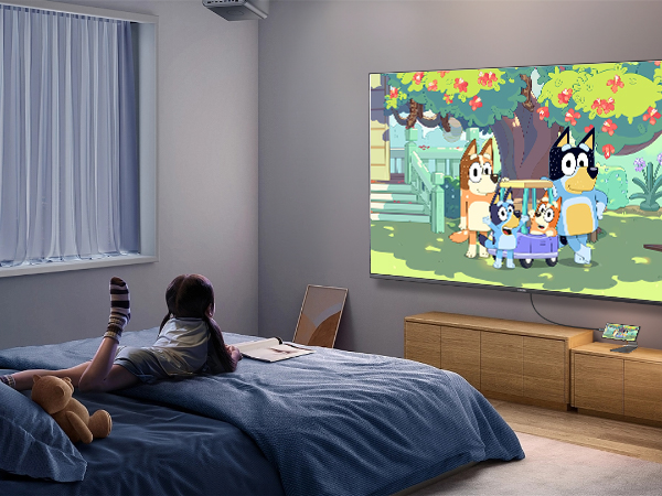 Home theater setup showing animated content projected on wall, with viewer silhouette on bed in darkened room.