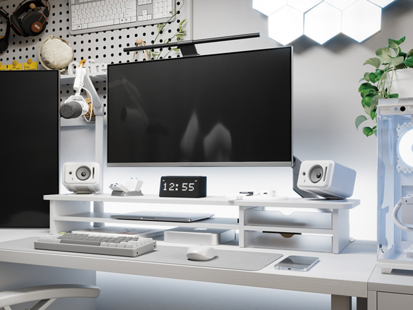 Modern desk setup with large monitor, white speakers, digital clock, keyboard, and decorative items on a white surface against a grey wall.