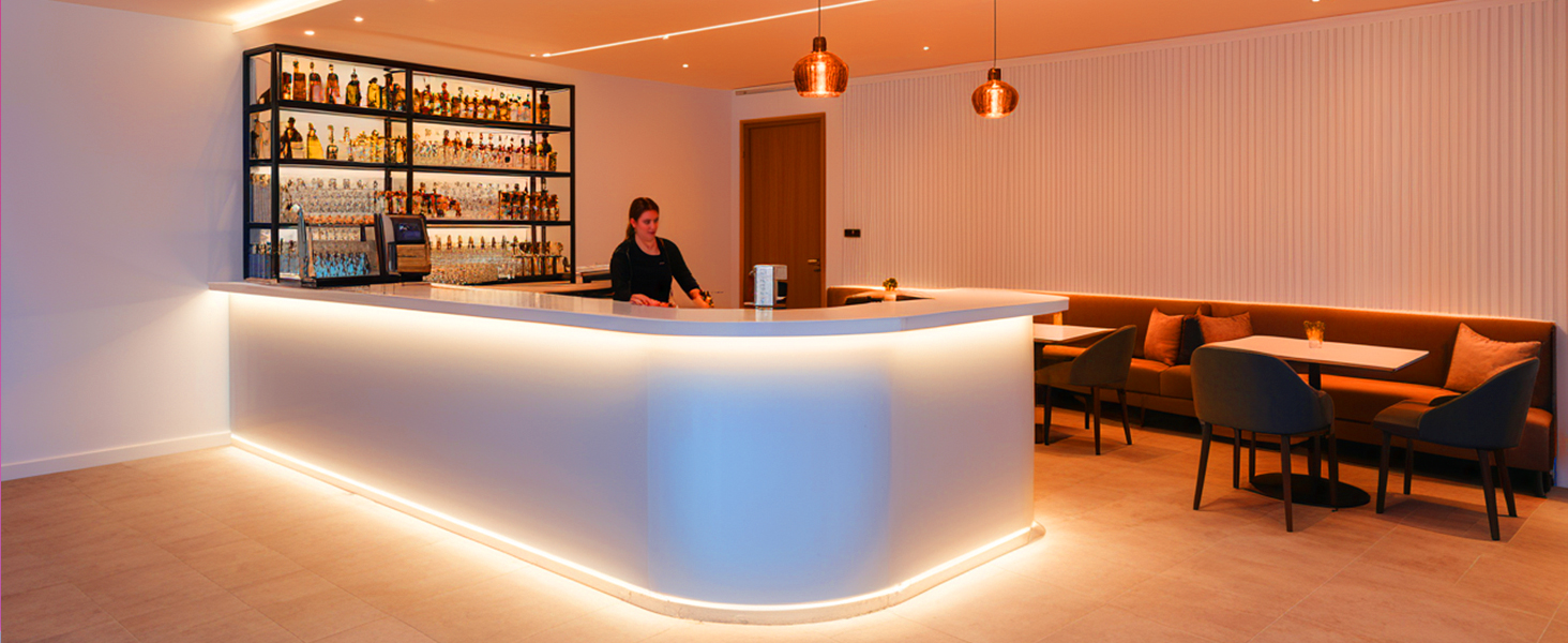 Modern bar interior with curved illuminated counter, featuring backlit shelving displaying bottles and ambient pendant lighting. Seating area with chairs visible in background.