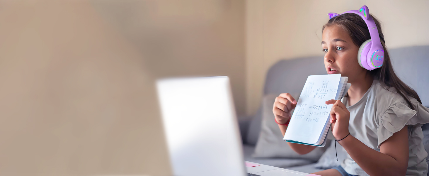 Wireless over-ear headphones with purple ear cups worn by a person holding a book while sitting in front of a laptop.