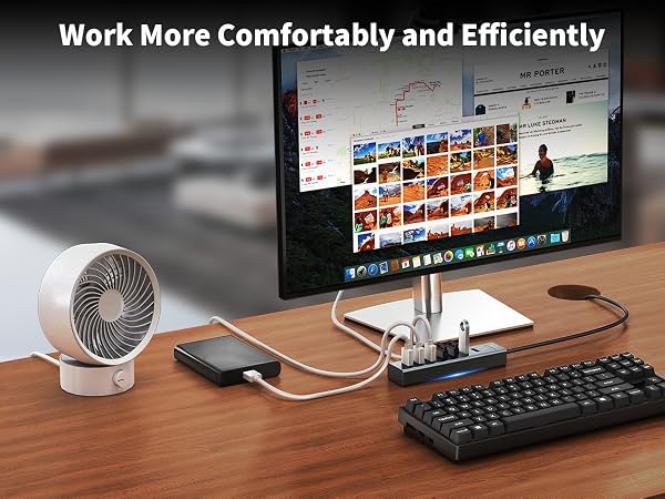 Desktop workstation setup with monitor, small portable fan, USB hub, and mechanical keyboard on wooden surface.