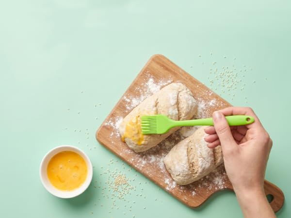 Green silicone brush applying egg wash on homemade bread dough with sesame on mint background
