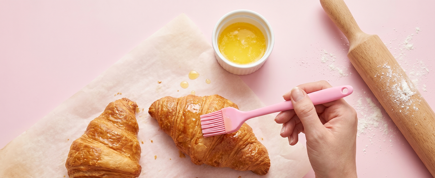Pink silicone pastry brush spreading melted butter on croissants on pastel pink background