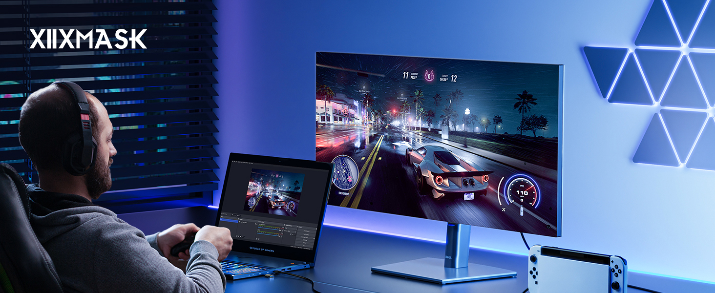 Gaming setup with large monitor displaying racing game, laptop, and person seated. Blue ambient lighting and triangular wall decorations. XIIXMASK text visible in upper left.