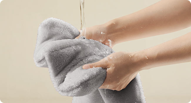 Hands washing a gray fluffy towel, creating soap suds and water droplets against a beige background.