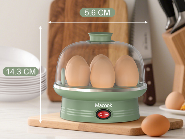 Macook egg cooker with clear dome lid, green base, and power button. Contains three eggs. Dimensions: 5.6 cm width, 14.3 cm height. Shown on kitchen counter with utensils.