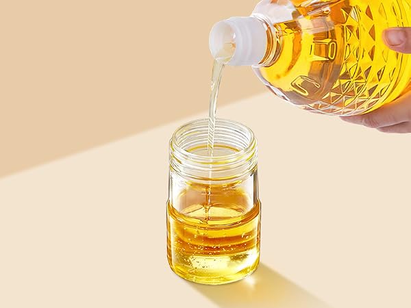 Yellow liquid being poured from a faceted glass bottle into a small clear jar. The liquid appears to be an oil or syrup.