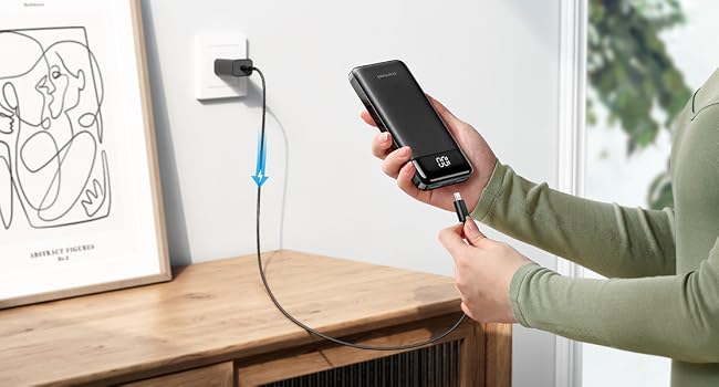 Person plugging smartphone into wall charger. Wooden surface with framed artwork visible in background.