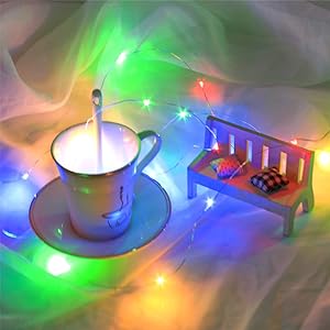 White teacup and saucer with steam rising, surrounded by colorful LED string lights creating a dreamy atmosphere with miniature bench.