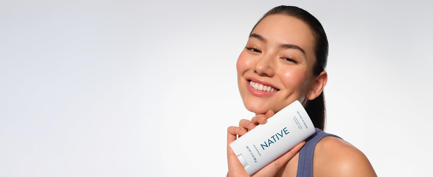 Smiling woman with fresh skin holds Native Sea Salt & Cedar deodorant, looking confident.