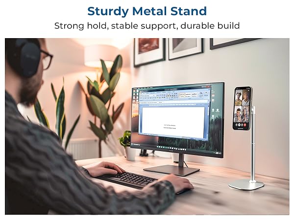 Metal phone stand on desk beside monitor and keyboard, with plant in background. Text reads 'Sturdy Metal Stand'.