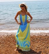 Long, flowy beach dress with blue and yellow swirling pattern. Sleeveless design worn by a person standing on a pebble beach with ocean in background.