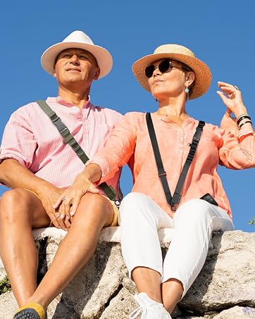 Two people in summer attire sit on a stone wall against a blue sky. They wear straw hats, pink shirts, and cross-body bags, suggesting outdoor leisure activity.