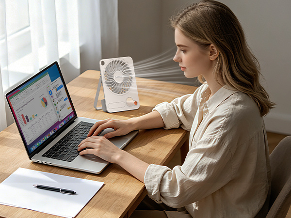 Woman working at wooden desk with laptop and water glass, wearing beige cardigan in bright natural light from window, depicting remote work or home office setup.