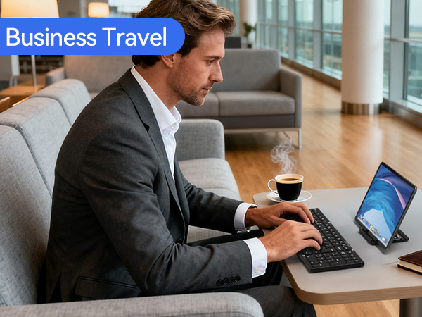 Text reads 'Business Travel'. Professional man in dark blazer seated on gray couch working on laptop with coffee cup nearby in modern office setting with city skyline view.