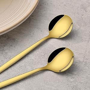 Two gold-plated serving spoons with long handles displayed on grey stone or concrete surface, showing elegant minimalist design.