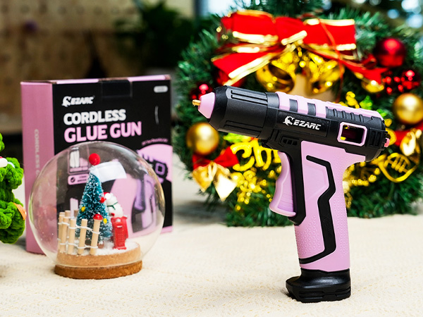 Pink and black cordless glue gun displayed with its retail packaging against festive Christmas decorations including bells and ornaments.