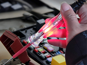 Close-up of a hand using a transparent electrical tester with glowing red light on a circuit board with colorful components.