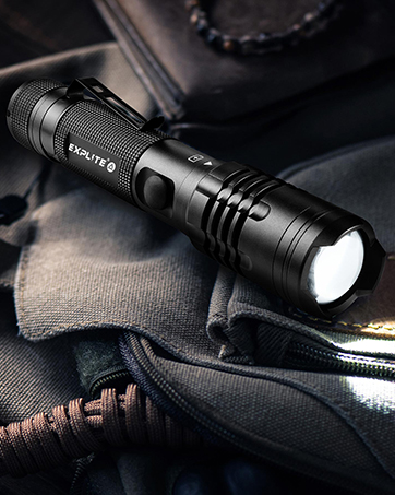Black tactical flashlight with ribbed grip and bright LED beam, shown on dark fabric with partial view of a bag strap.
