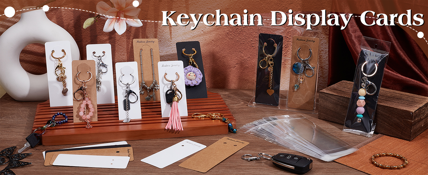 Display of various keychain cards with attached decorative charms and tassels. Wooden stand holds multiple cards showing different designs. Packaging materials and accessories visible in background.