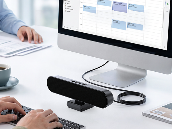 Compact black microphone with cable attached next to computer monitor displaying calendar application on white desk with wireless mouse and keyboard.