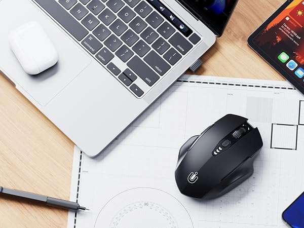 Desktop workspace setup showing a wireless mouse, laptop, mobile device, and writing materials arranged on a wooden surface.