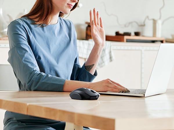 Person in blue shirt sitting at desk with laptop, holding hand up as if waving or gesturing during video call.