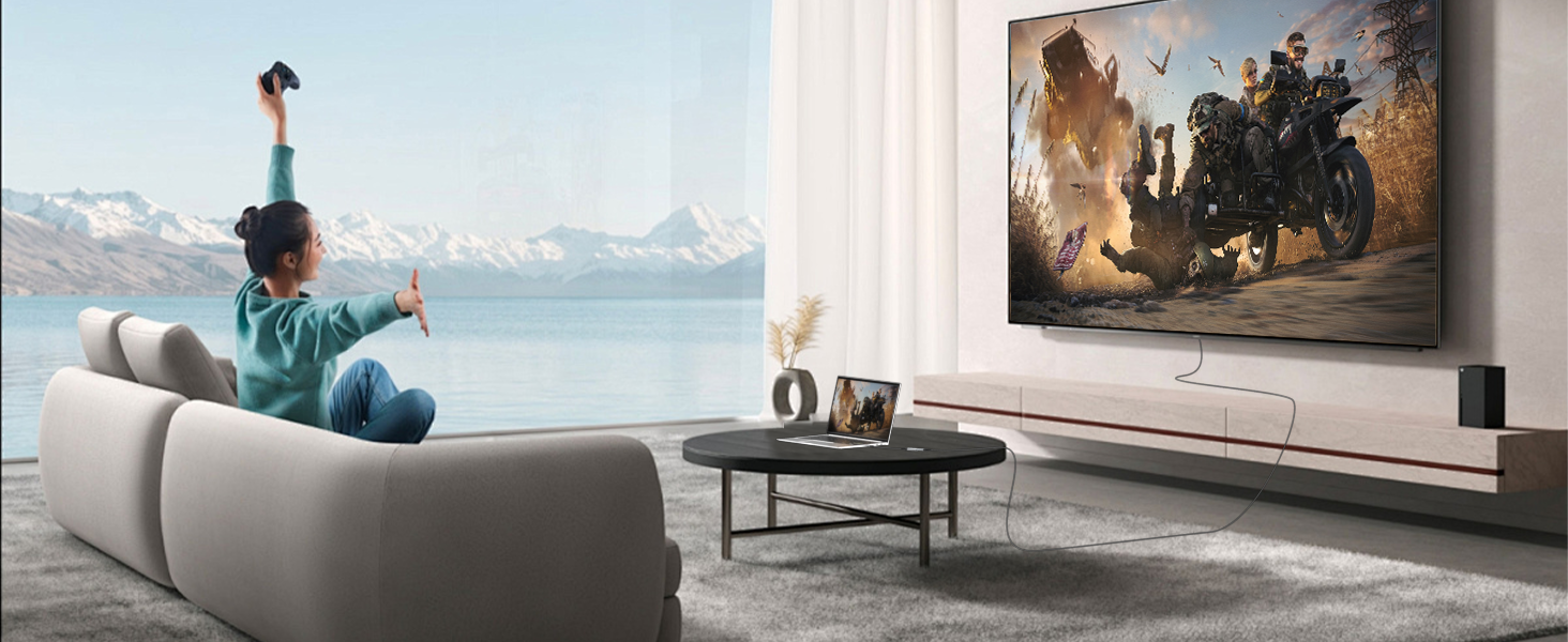 Modern living room setup showing a large wall-mounted TV display with soundbar, featuring a gray sofa and coffee table with mountain view through windows.