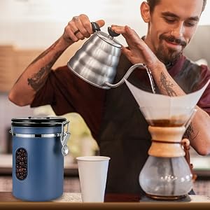 Person pouring hot water from a gooseneck kettle into a pour-over coffee maker. Blue canister with viewing window visible on counter.