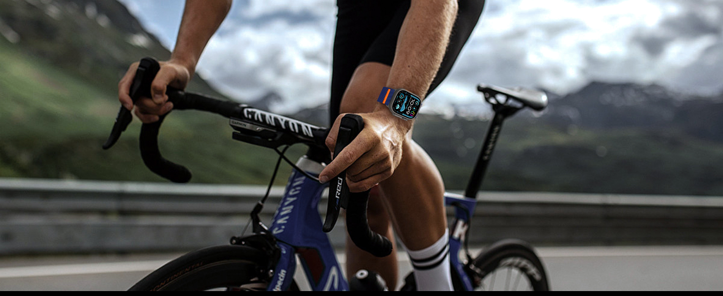 Cyclist's hands gripping handlebars of a road bike, wearing a digital watch. Mountain scenery visible in the background.