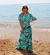 Woman wearing long, flowy teal dress with floral pattern, standing on beach with arms akimbo. Sun hat and ocean visible in background.